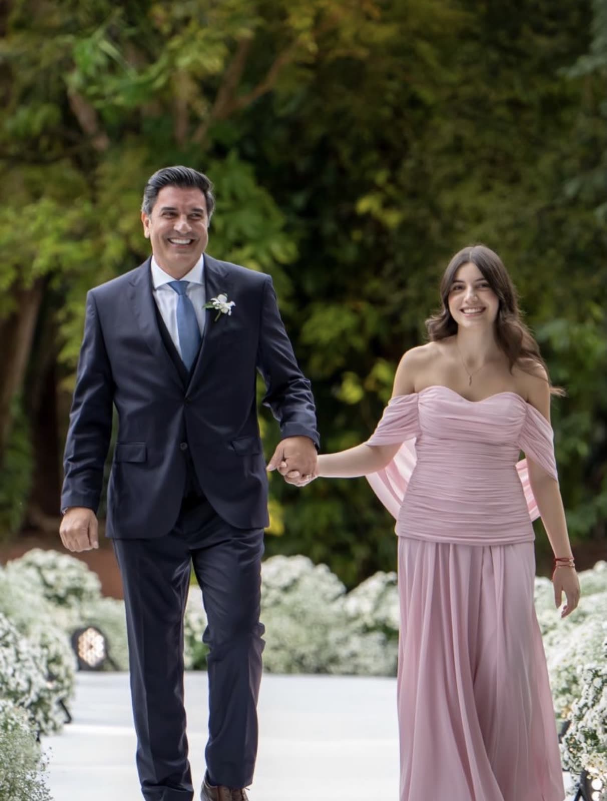 Pai e filha de mãos dadas, caminhando pelo tapete branco do jardim