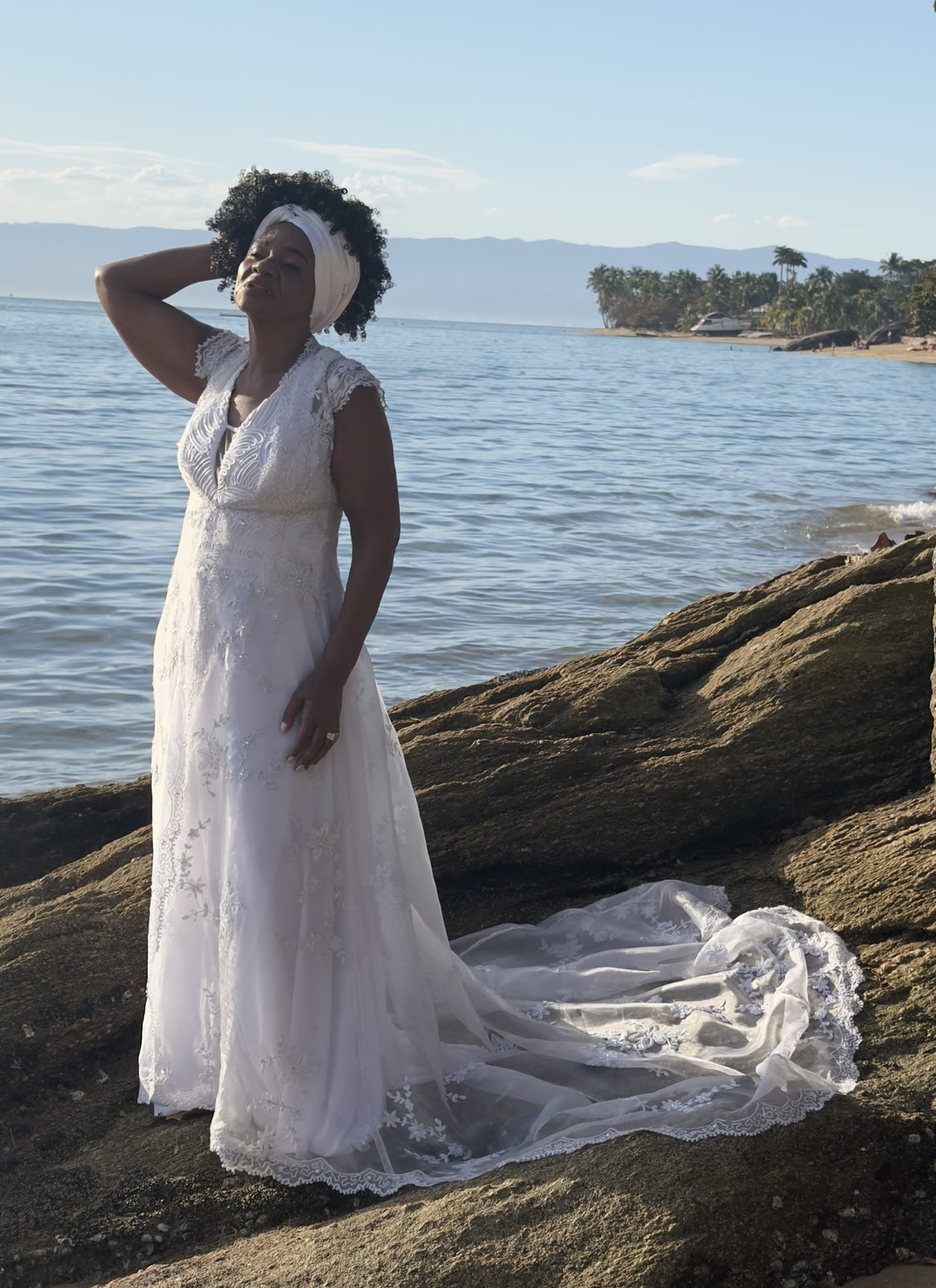 Noiva na beira do mar, rosto erguido ao sol, vestido de renda ao vento